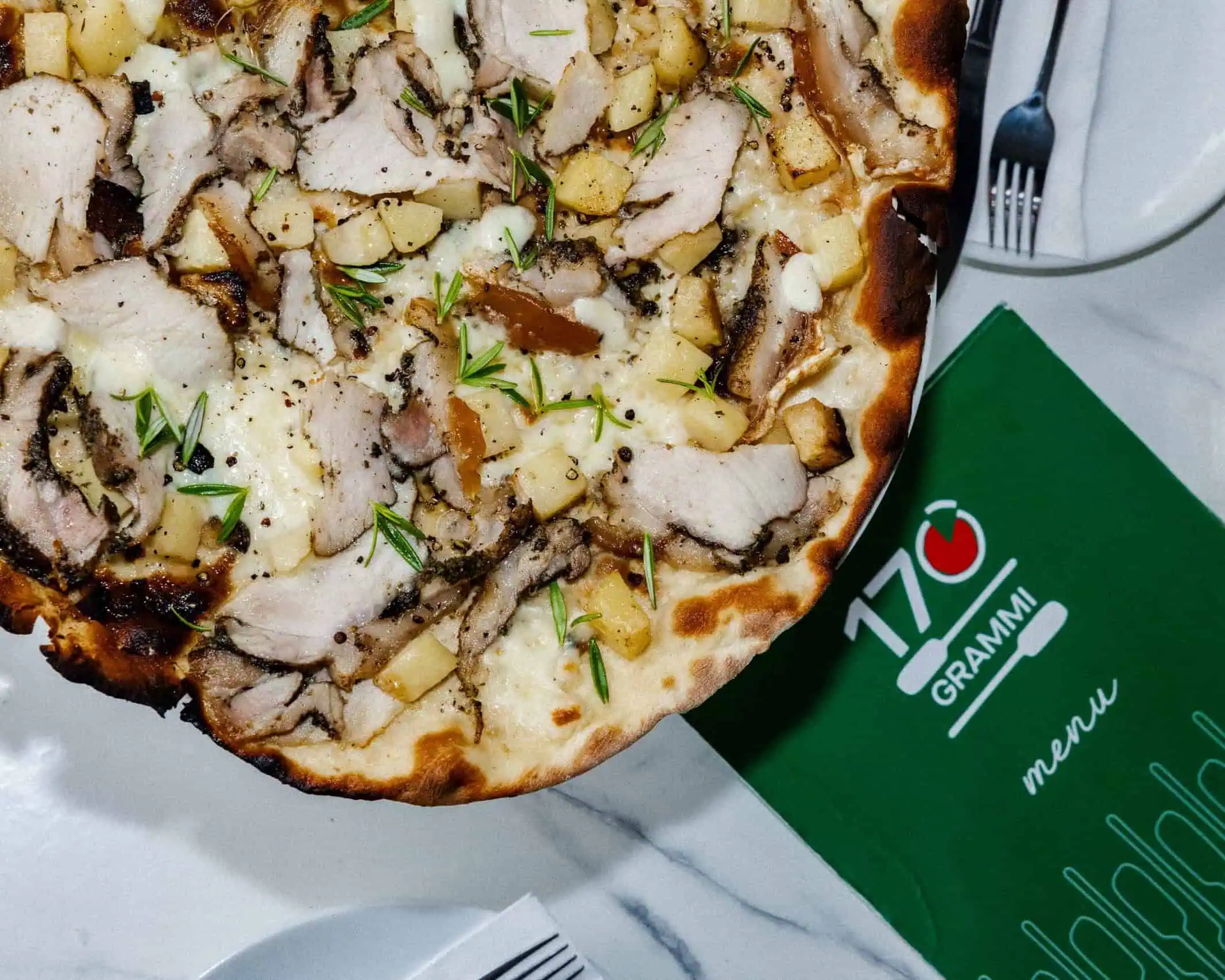 Close-up of a 170 Grammi Roman-style white pizza topped with melted mozzarella, sliced porchetta, diced potato, cracked pepper, and fresh rosemary on a restaurant table.