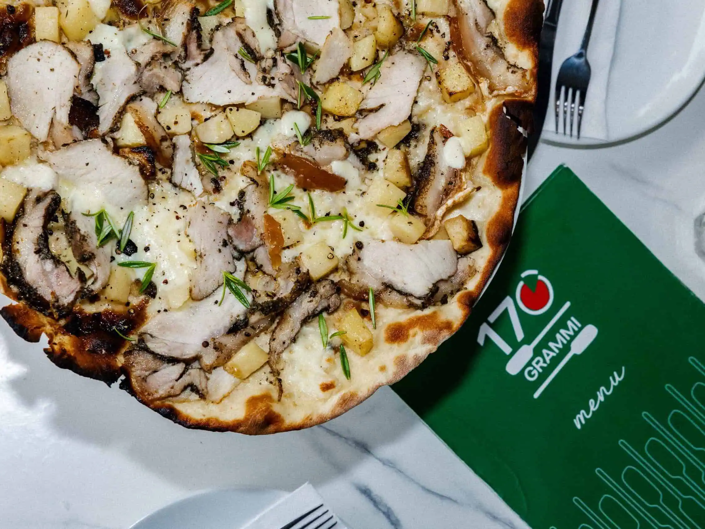 Close-up of a 170 Grammi Roman-style white pizza topped with melted mozzarella, sliced porchetta, diced potato, cracked pepper, and fresh rosemary on a restaurant table.
