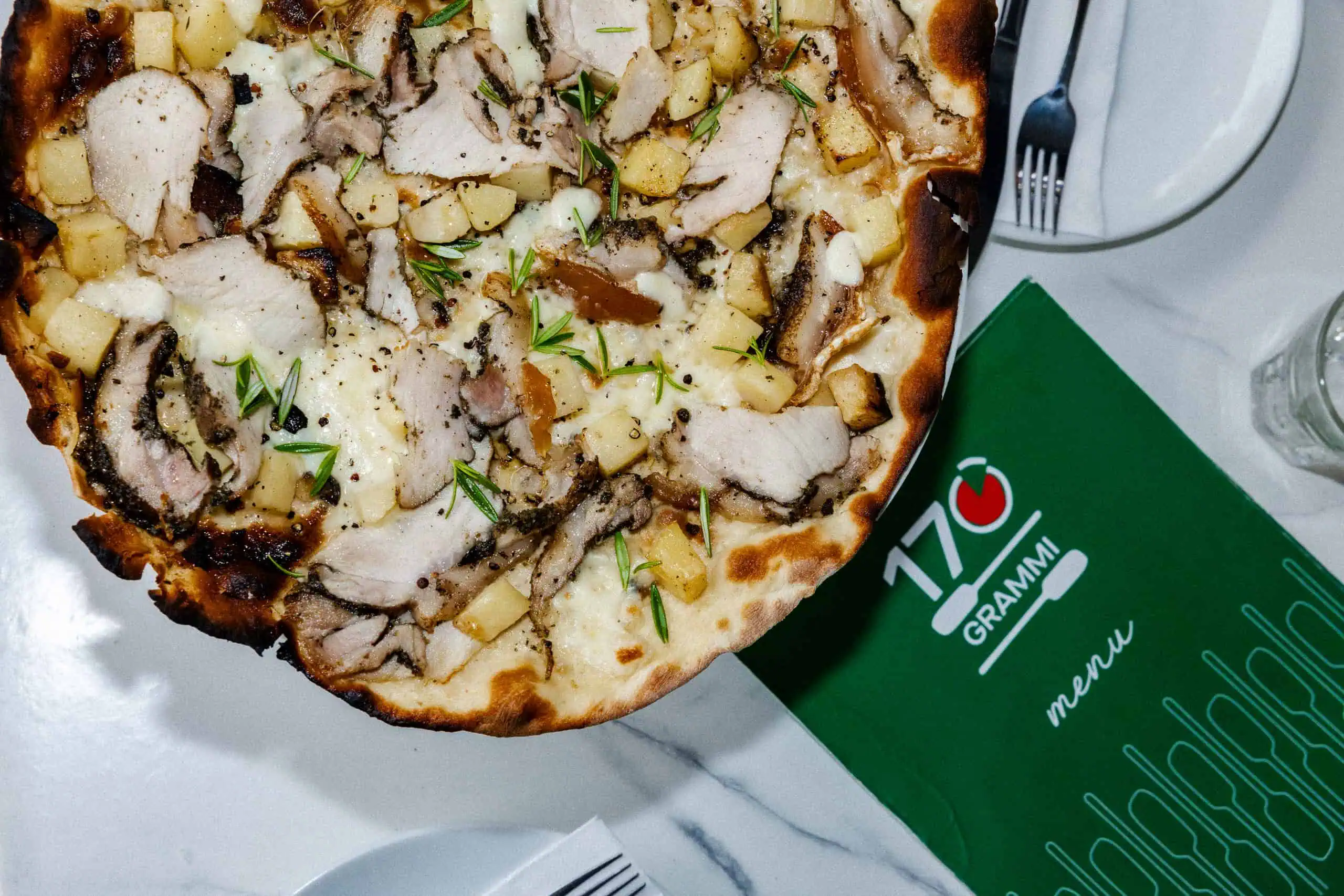 Close-up of a 170 Grammi Roman-style white pizza topped with melted mozzarella, sliced porchetta, diced potato, cracked pepper, and fresh rosemary on a restaurant table.