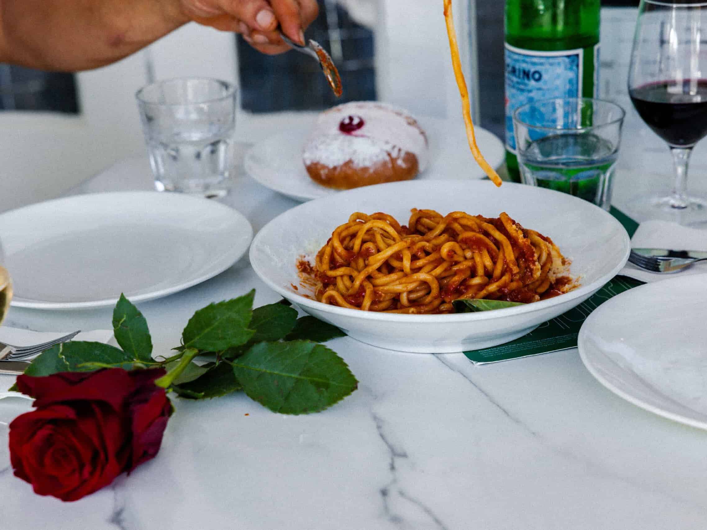 owl of spaghetti in tomato sauce on a white restaurant table with wine, water and a powdered-sugar dessert, with a hand twirling pasta at 170 Grammi.