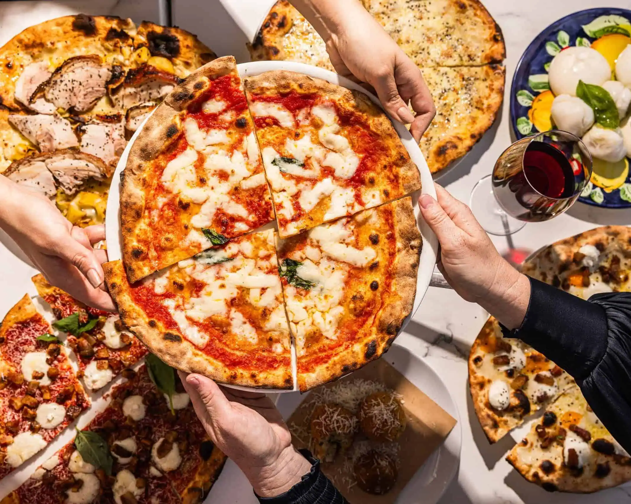 Hands sharing a variety of wood-fired Italian pizzas including Margherita, porchetta, and spicy salame, with burrata and wine at 170 Grammi in Sydney