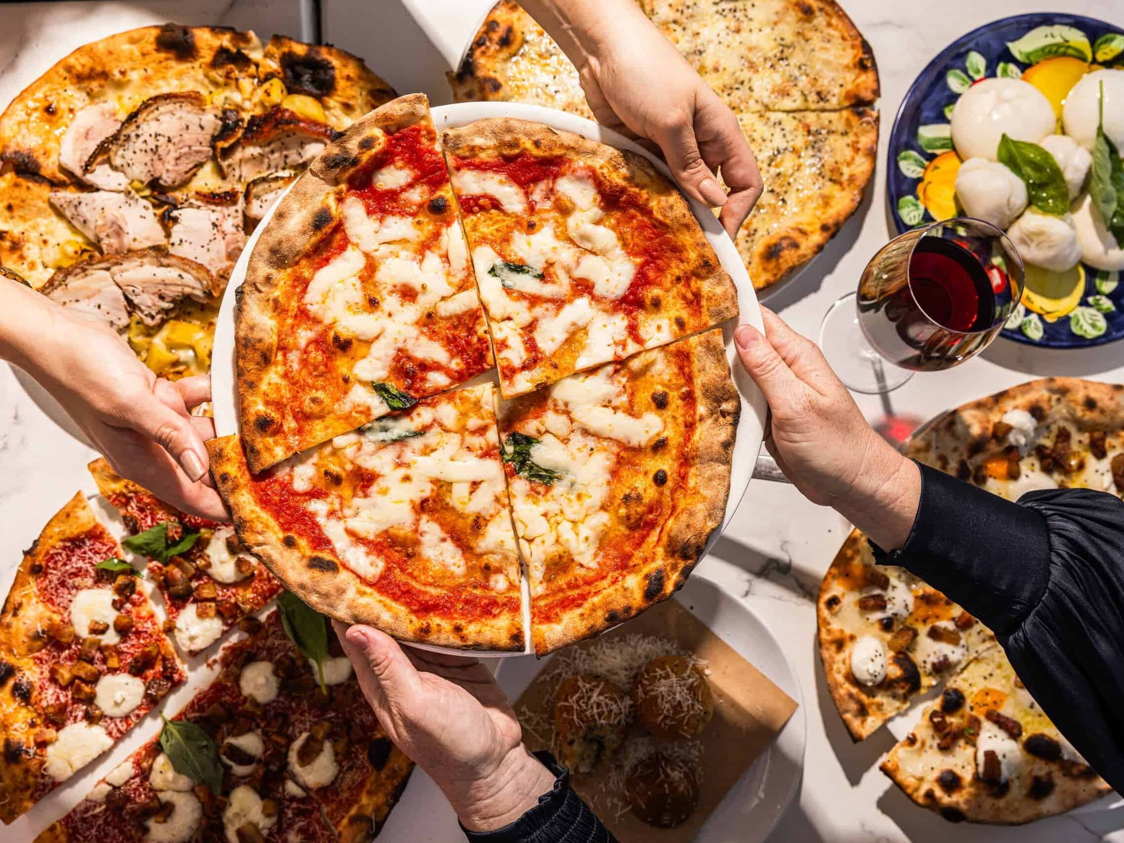 Hands sharing a variety of wood-fired Italian pizzas including Margherita, porchetta, and spicy salame, with burrata and wine at 170 Grammi in Sydney