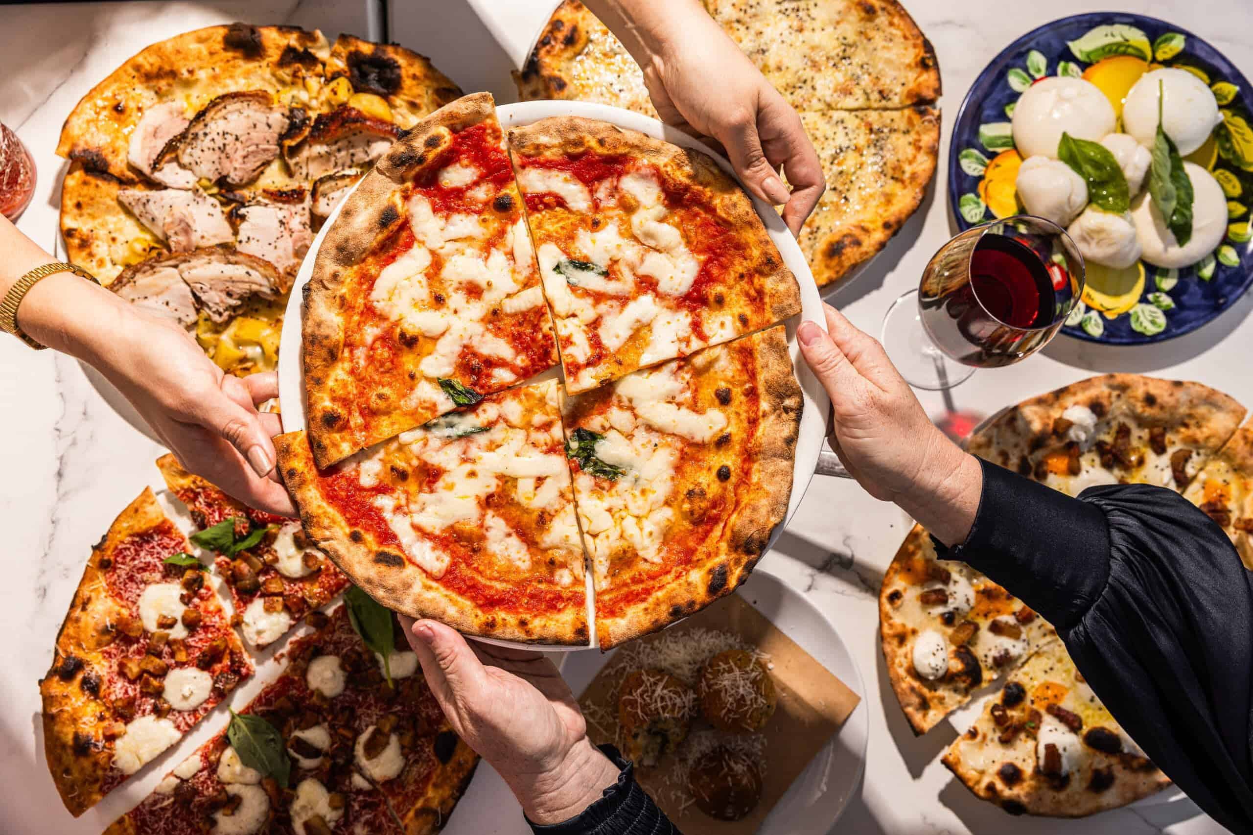 Hands sharing a variety of wood-fired Italian pizzas including Margherita, porchetta, and spicy salame, with burrata and wine at 170 Grammi in Sydney