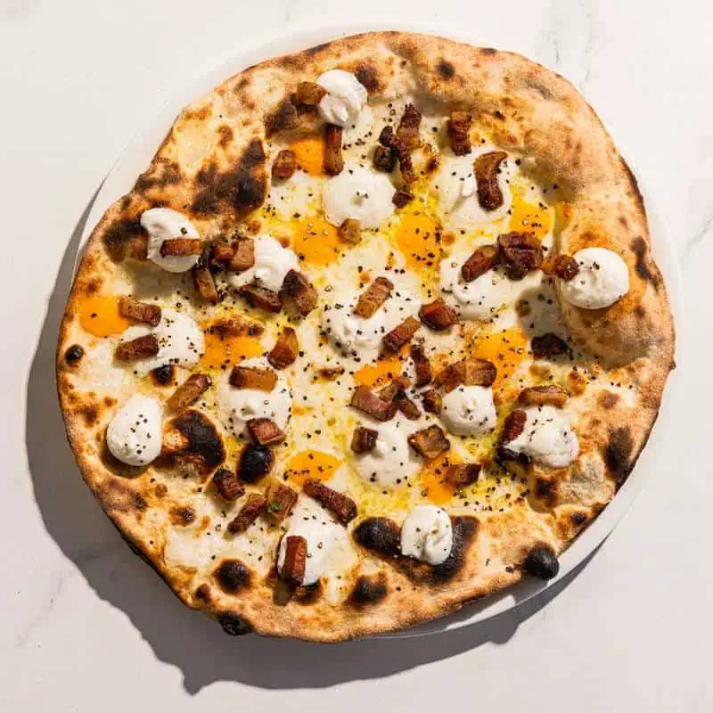 Carbonara-inspired pizza with a white base, topped with Pecorino Romano, guanciale, free-range egg yolk, and black pepper