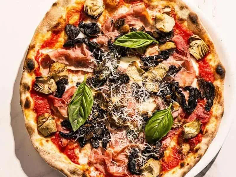 Capricciosa pizza topped with San Marzano tomato, buffalo mozzarella, double smoked ham, sautéed mushrooms, marinated artichokes, and fresh basil