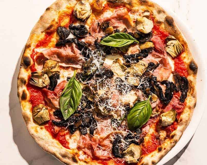Capricciosa pizza topped with San Marzano tomato, buffalo mozzarella, double smoked ham, sautéed mushrooms, marinated artichokes, and fresh basil