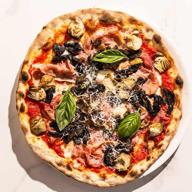 Capricciosa pizza topped with San Marzano tomato, buffalo mozzarella, double smoked ham, sautéed mushrooms, marinated artichokes, and fresh basil