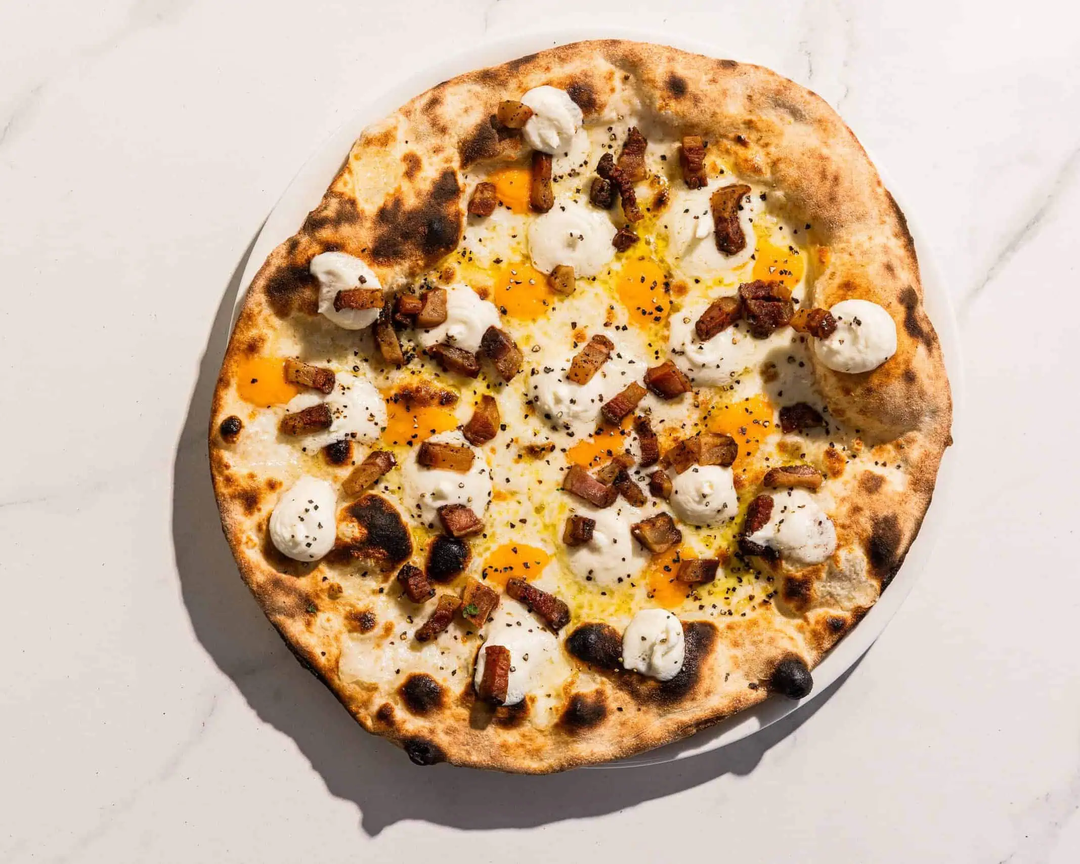 Carbonara-inspired pizza with a white base, topped with Pecorino Romano, guanciale, free-range egg yolk, and black pepper