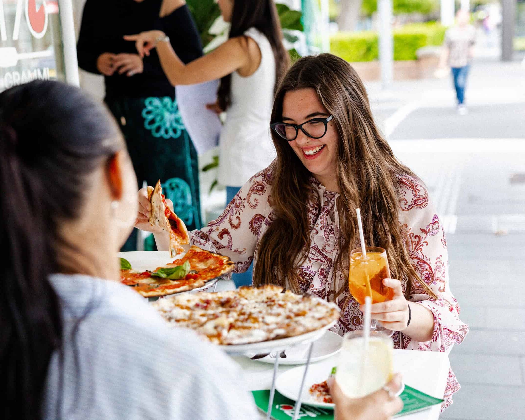 Smiling woman enjoying the La Dolce Vita lunch special at 170 Grammi, lifting a slice of pizza and holding an iced drink at an outdoor table.