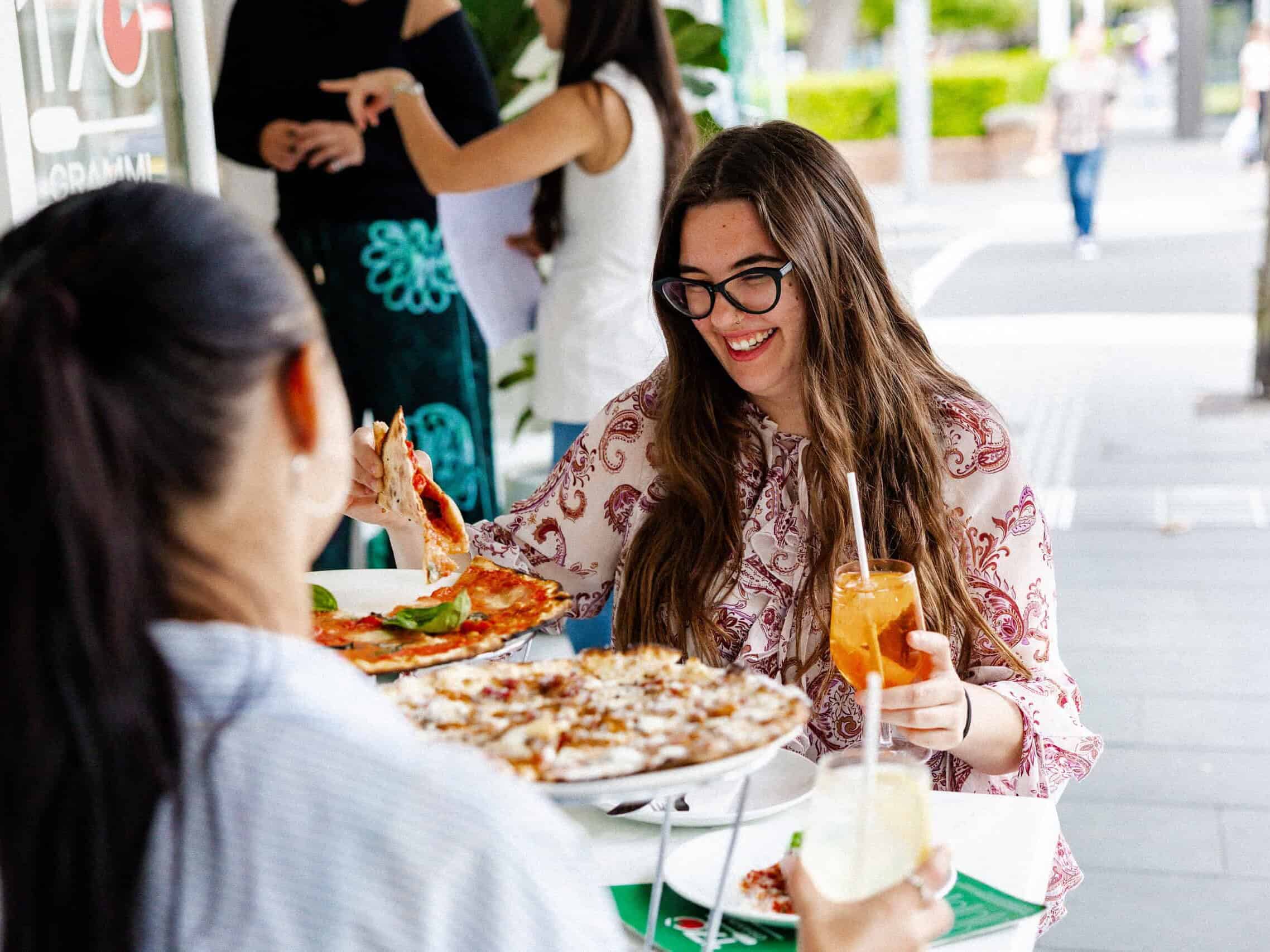 Smiling woman enjoying the La Dolce Vita lunch special at 170 Grammi, lifting a slice of pizza and holding an iced drink at an outdoor table.