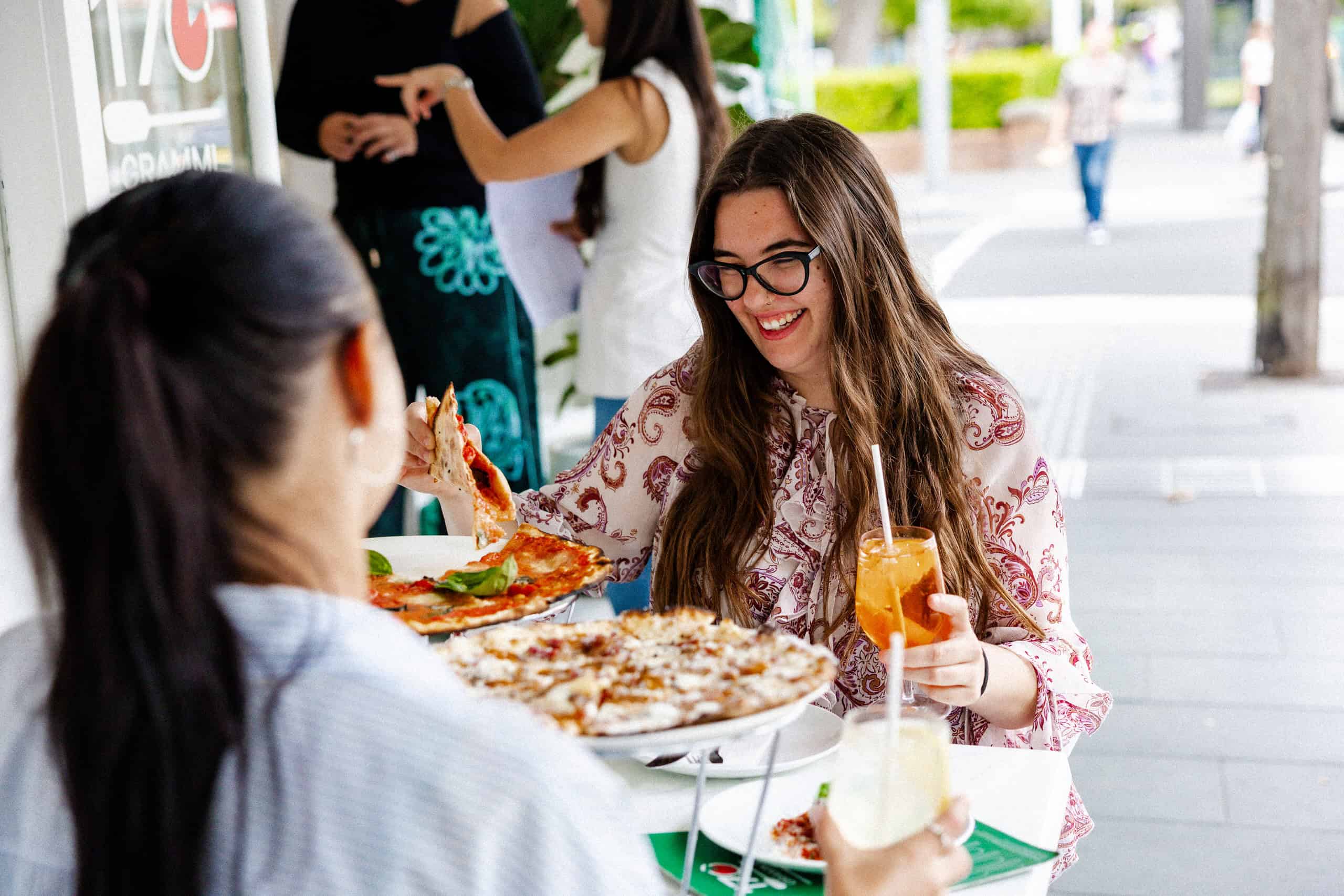 Smiling woman enjoying the La Dolce Vita lunch special at 170 Grammi, lifting a slice of pizza and holding an iced drink at an outdoor table.