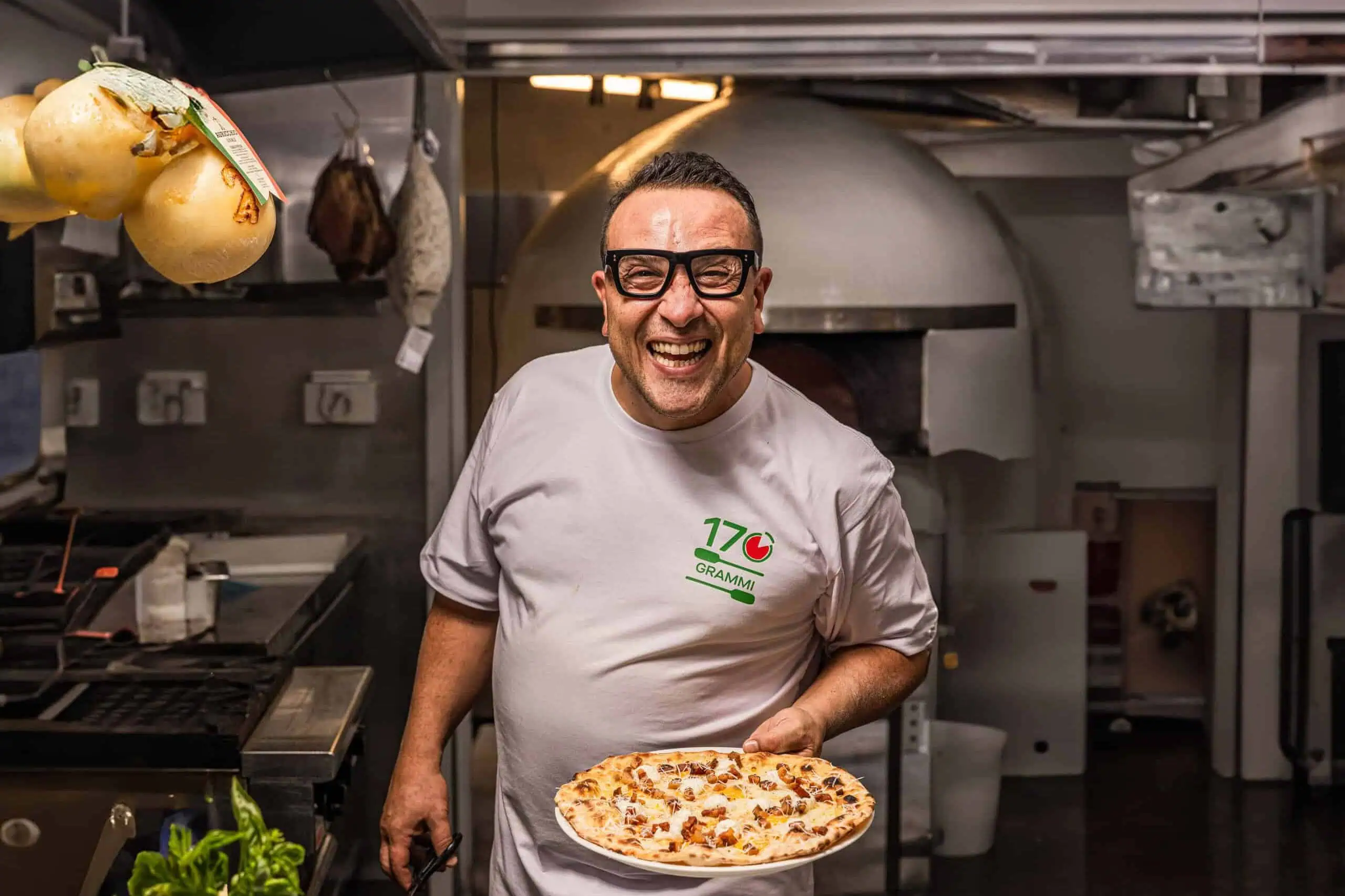 Smiling pizzaiolo, Luigi Espositio, in the kitchen at 170 Grammi holding a freshly prepared wood-fired pizza in front of the pizza oven
